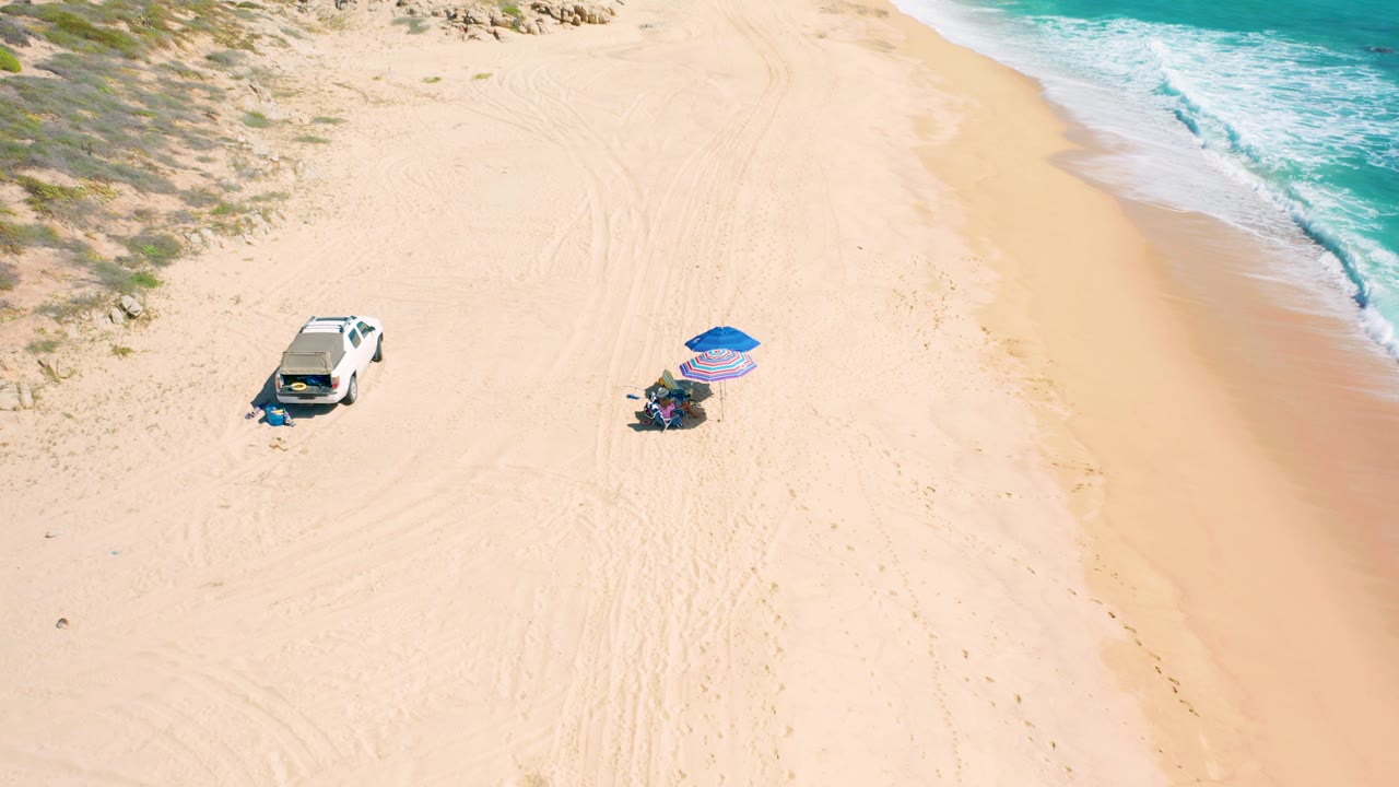 High aerial frame over the El Cardoncito beach showing the parked white car, a blue umbrella with a striped shade, small visible people, tire tracks in the sand, and waves breaking near the upper right corner.