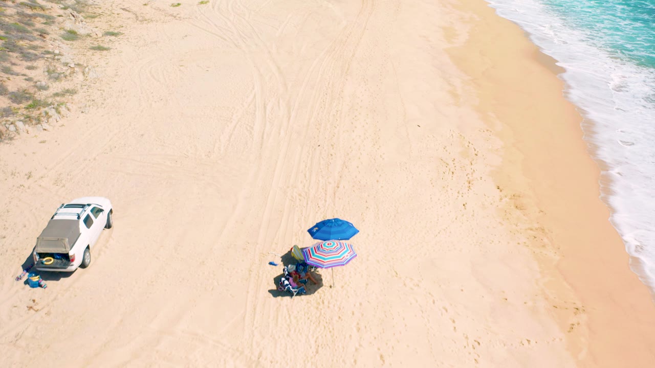 High aerial frame over El Cardoncito beach with a white SUV on the left, blue and striped umbrellas on the sand, small people visible, and turquoise surf along the right edge.