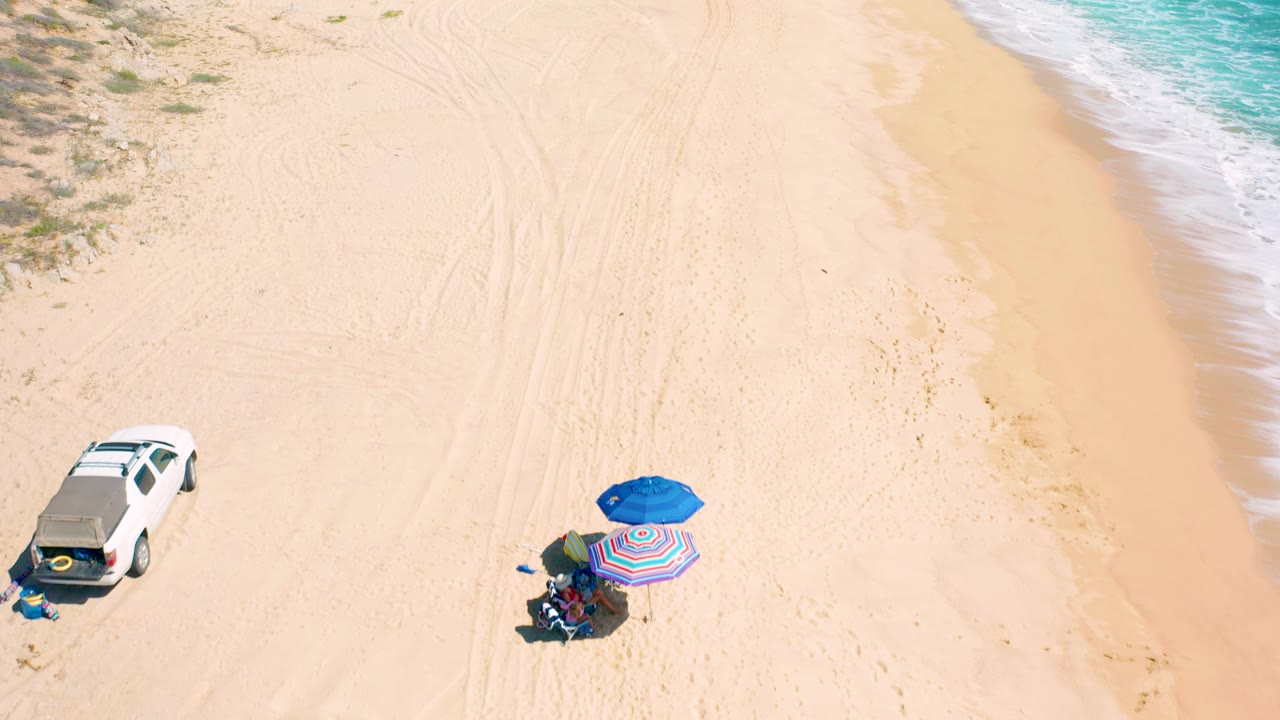 High overhead frame in El Cardoncito showing the white SUV closer to the lower left, the blue and striped umbrellas to the lower right, people beneath them, and surf receding at the far right.