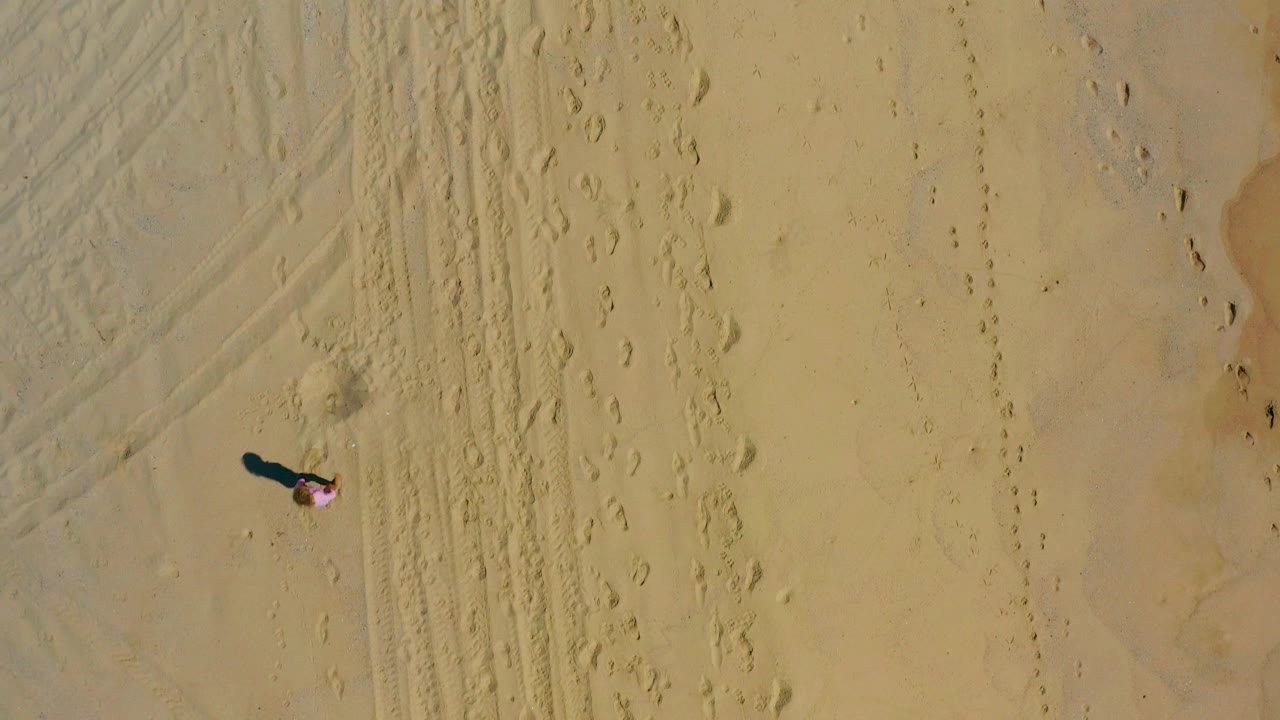 Near-vertical aerial frame from El Cardoncito, Baja California Sur, showing a small hat-wearing person on tan sand beside footprints, tire-like tracks, and a dark shadow.