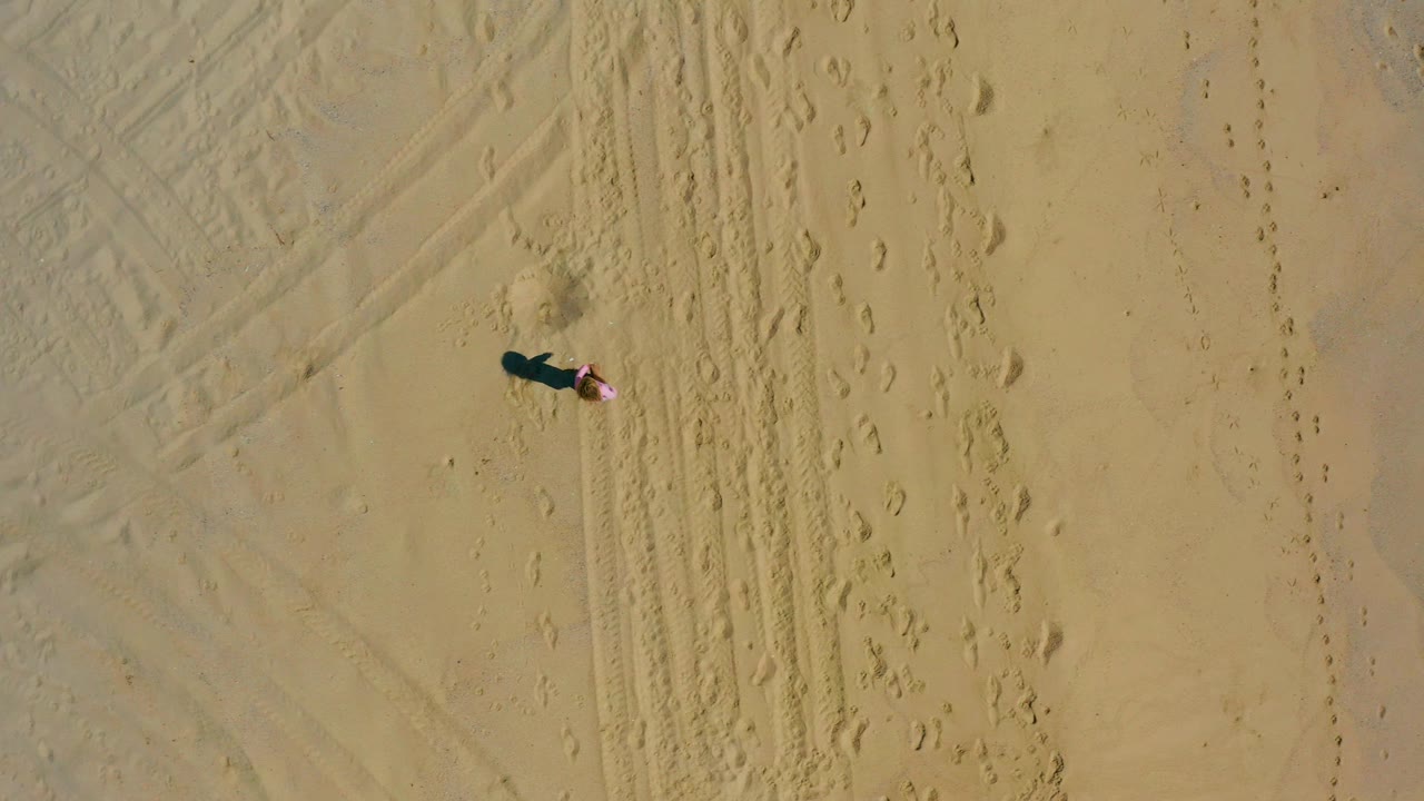Aerial frame from El Cardoncito with the person slightly lower in the sandy field, beside parallel tire tracks and scattered footprints.