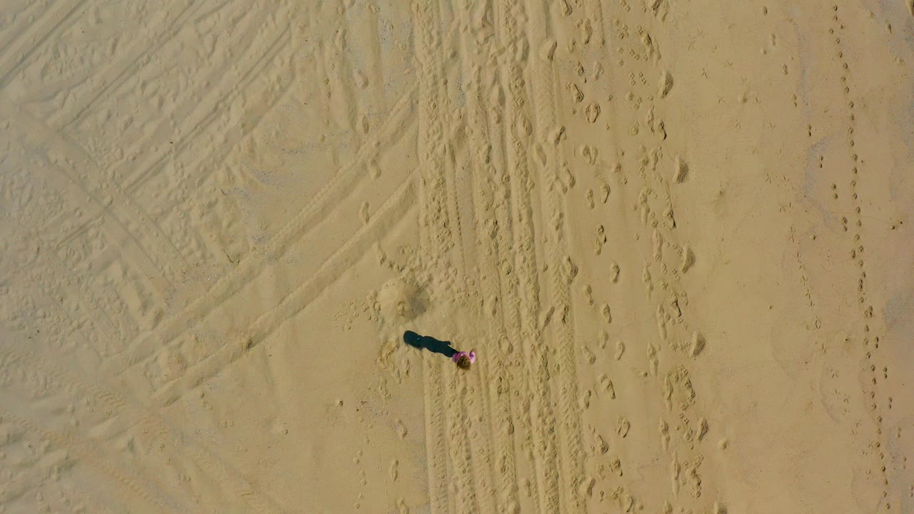 Overhead frame in El Cardoncito, Baja California Sur showing a small person in pink on pale sand beside tire tracks and footprints.