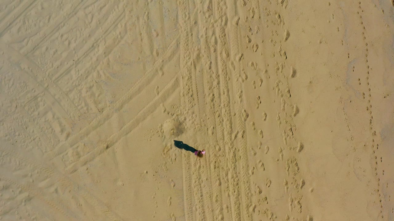 Overhead drone-style frame in El Cardoncito, Baja California Sur showing the runner in pink on the left side of marked sand with footprints to the right.