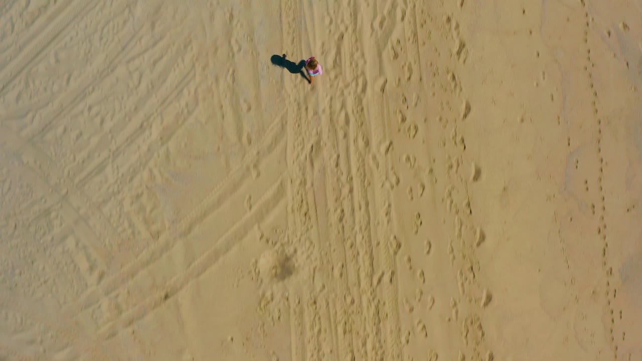 Overhead view in El Cardoncito, Baja California Sur showing a small child in pink near the upper frame on tan sand with footprints, track marks, and a long shadow.