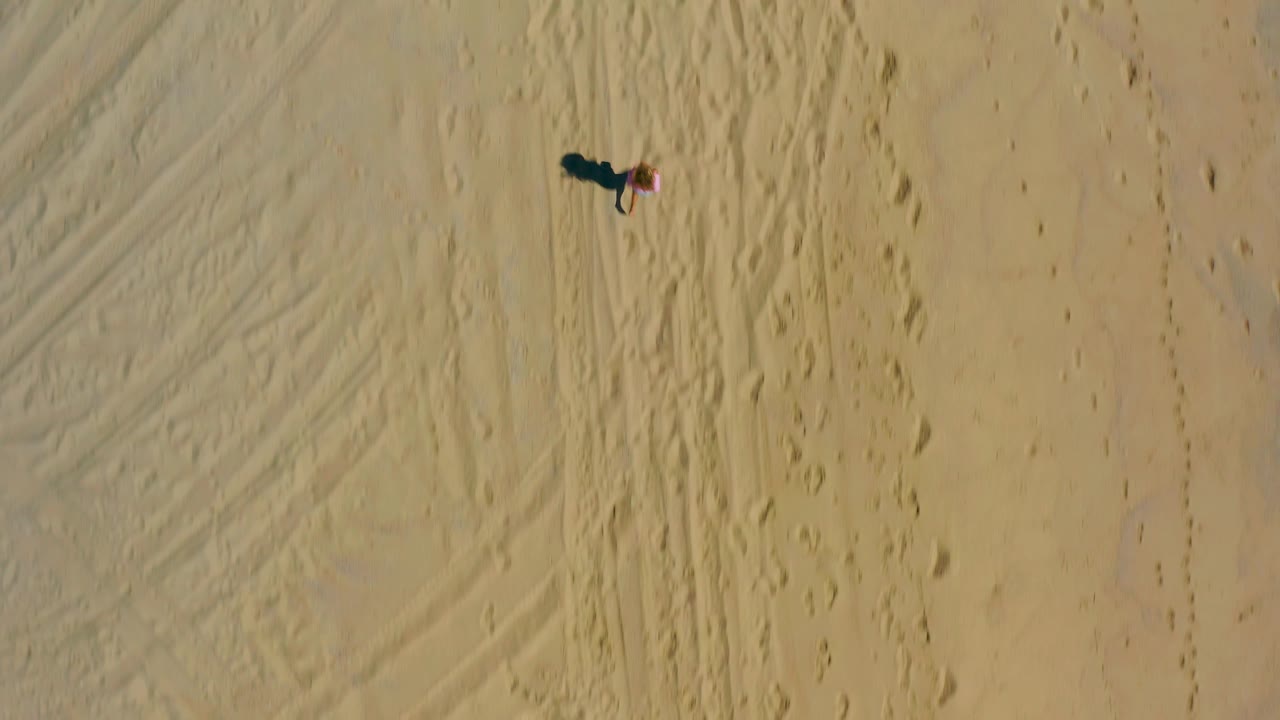 Aerial frame in El Cardoncito with the child near the upper right edge, surrounded by broad tan sand, foot trails, and faint compressed lines in the beach surface.