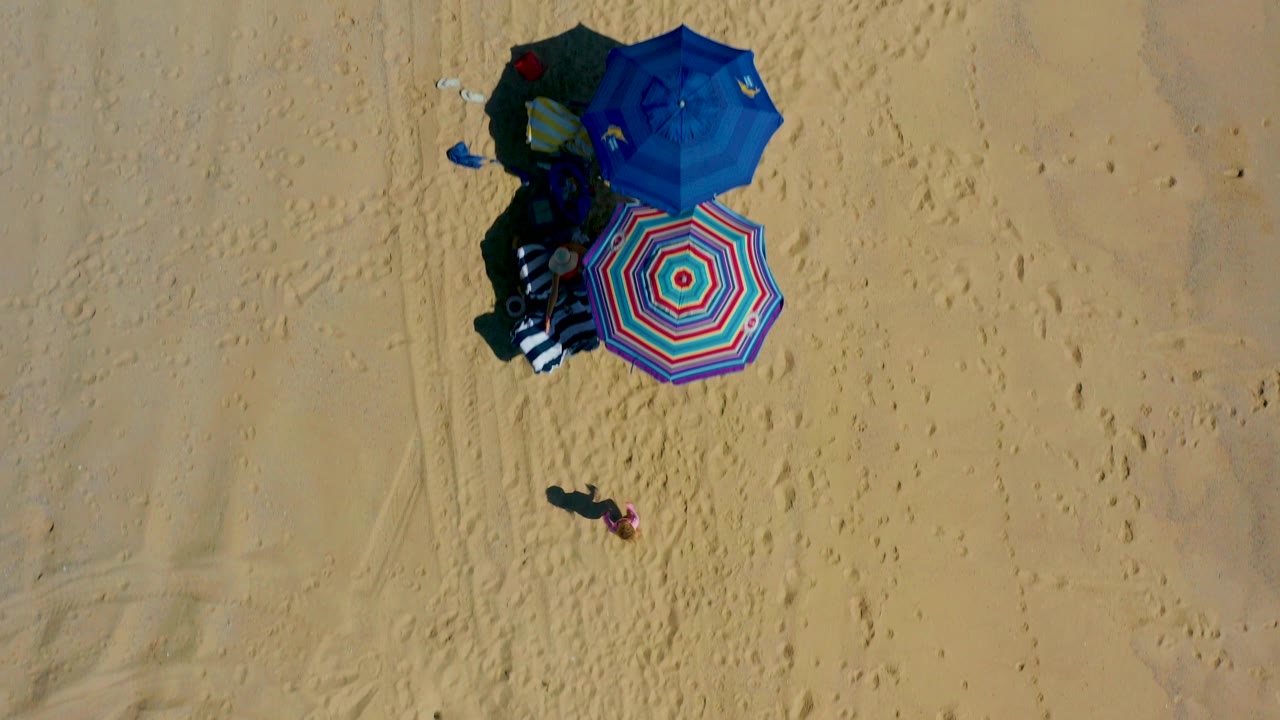 High overhead view in El Cardoncito, Baja California Sur, with blue and striped umbrellas clustered on sand and a small visible person standing below them.