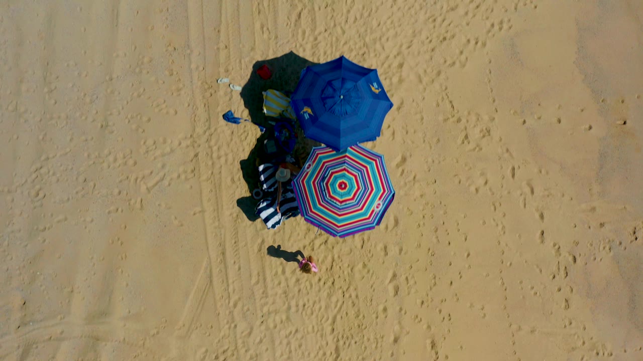 Top-down El Cardoncito beach view with two colorful umbrellas, dark shadows, packed belongings, and textured sand with footprints to the right.