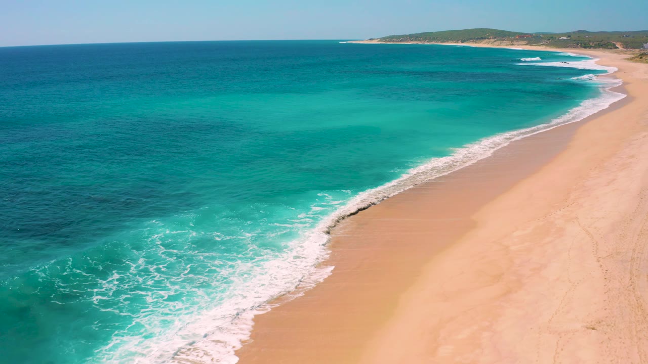 High oblique view of the Baja California Sur coast with darker blue ocean offshore, lighter water near the surf, a smooth sandy beach, and no visible people.