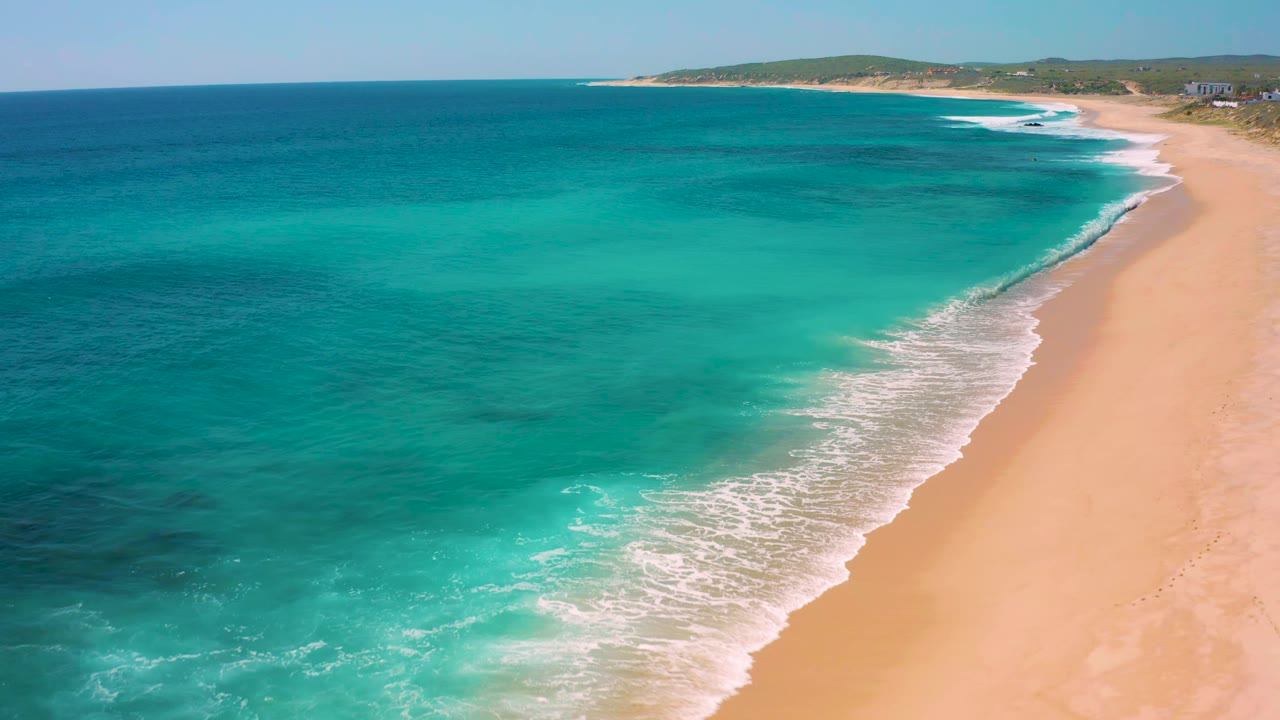 Drone view in El Cardoncito with blue-green ocean filling most of the frame, white foam tracing the beach edge, golden sand to the right, low coastal hills in back, and no visible people.