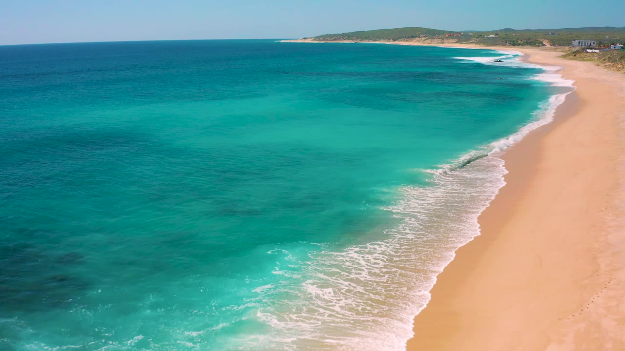 Aerial frame over the El Cardoncito shoreline where teal shallows meet deeper blue ocean, white wave foam cuts diagonally across the sand, and no visible people appear on the beach.