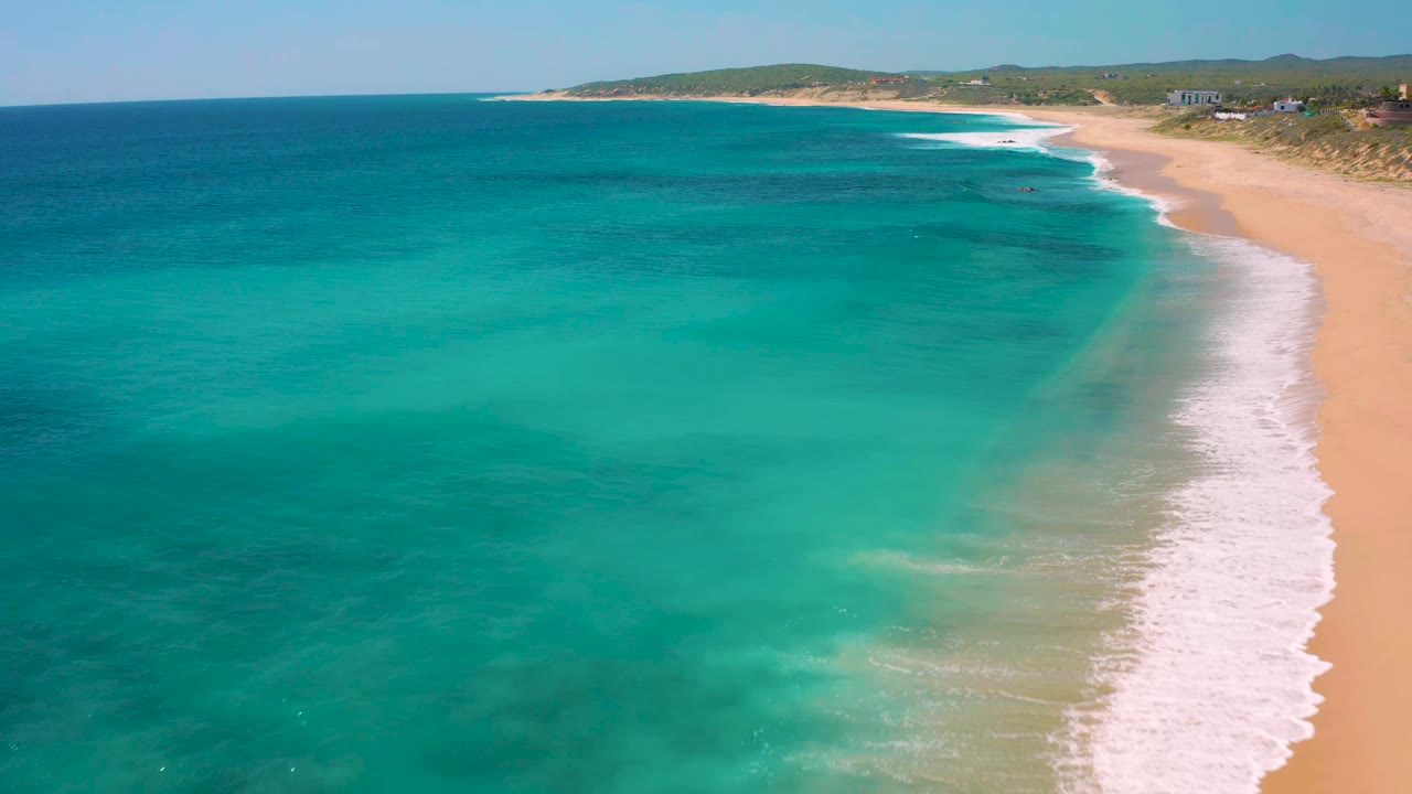 El Cardoncito coastline seen from above, with shallow turquoise water in the foreground, a narrow white wave band along the right edge, beach and dunes beyond it, and no visible people.