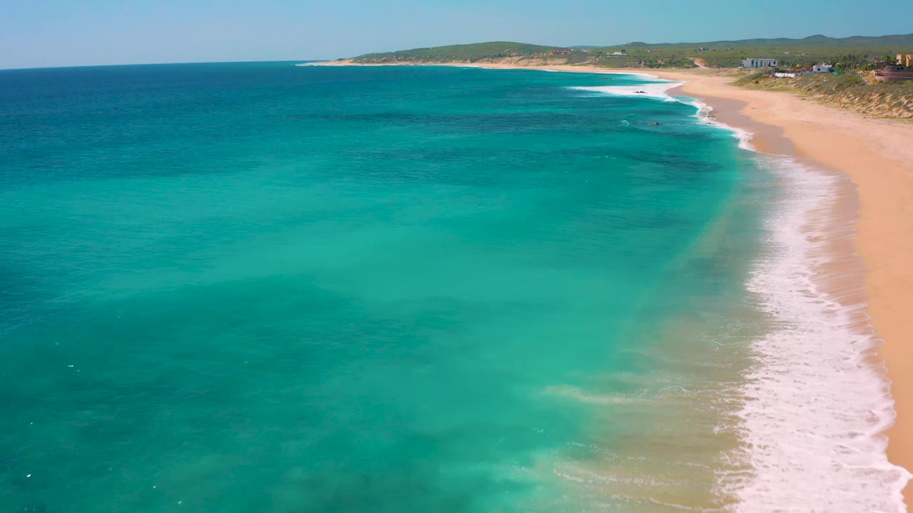 High aerial frame of El Cardoncito with blue-green water dominating the view, the sandy beach running diagonally on the right, low dunes and small coastal structures in the distance, and no visible people.