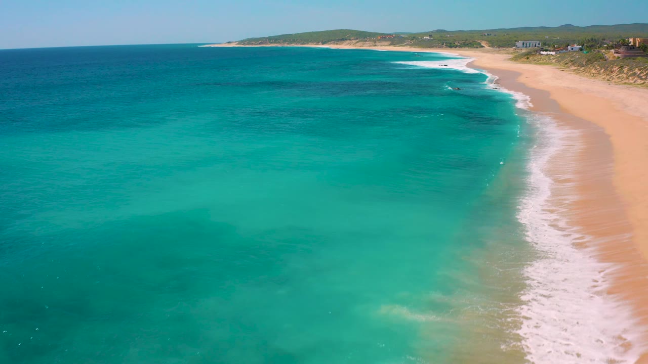 Aerial beach view with turquoise water, surf, sand, dunes, and distant hills.