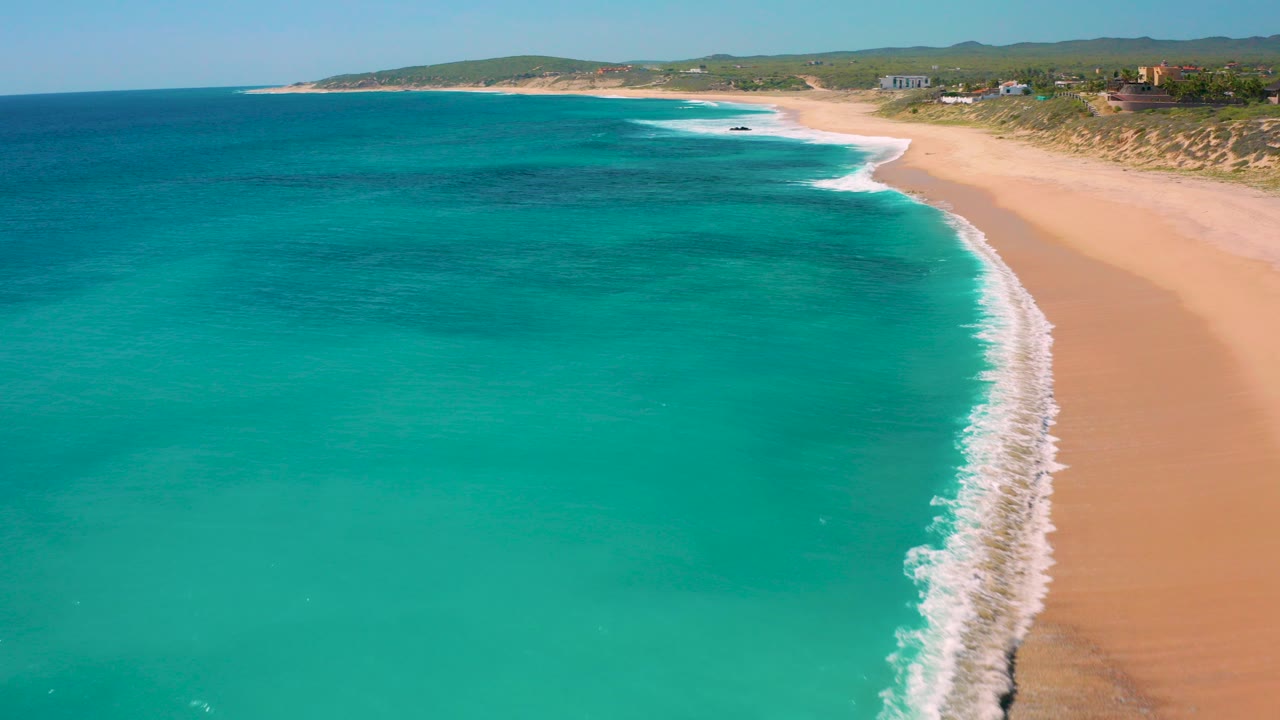 Aerial frame over El Cardoncito, Baja California Sur showing turquoise water, a diagonal white surf line, tan sand, low dunes, and no visible people.