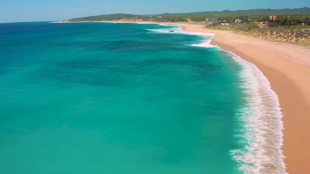 El Cardoncito aerial frame with the breaking wave line tight to the right edge, broad blue-green water across the foreground, tan beach beyond it, and no visible people.