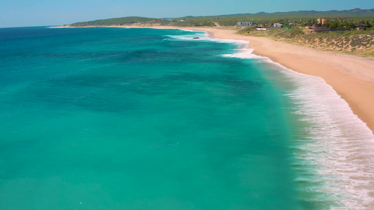 A wider El Cardoncito shoreline frame shows darker patches in the water, white surf bands, a long empty beach, low dune vegetation, and no visible people.
