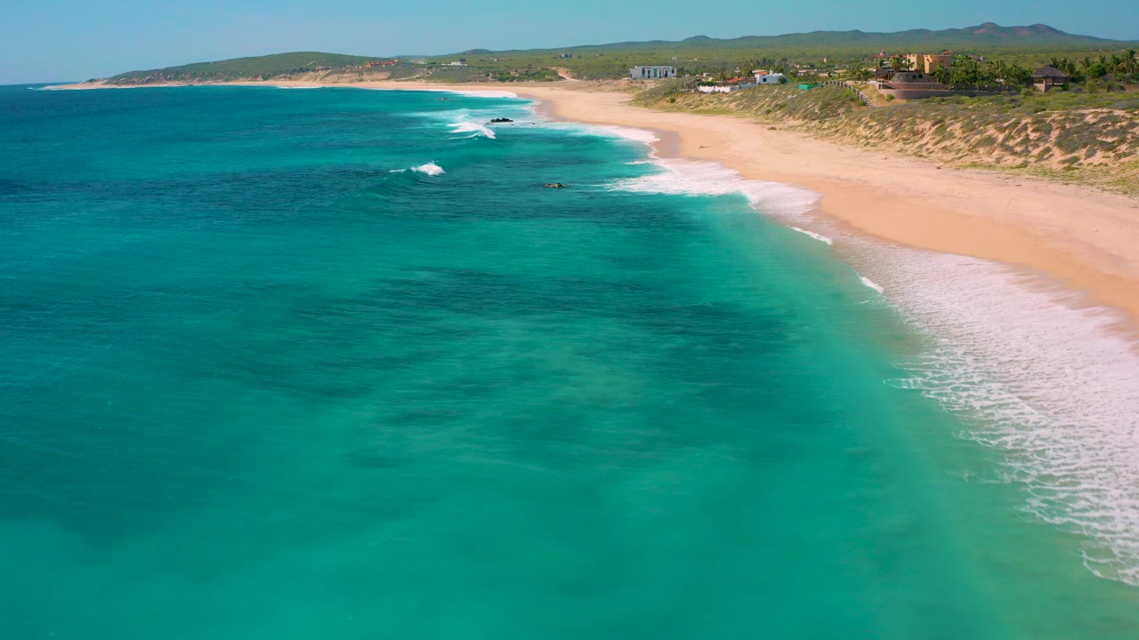 Aerial view over El Cardoncito, Baja California Sur, with turquoise water in the foreground, a pale sandy shoreline, scattered beachfront homes beyond the dunes, and no visible people.