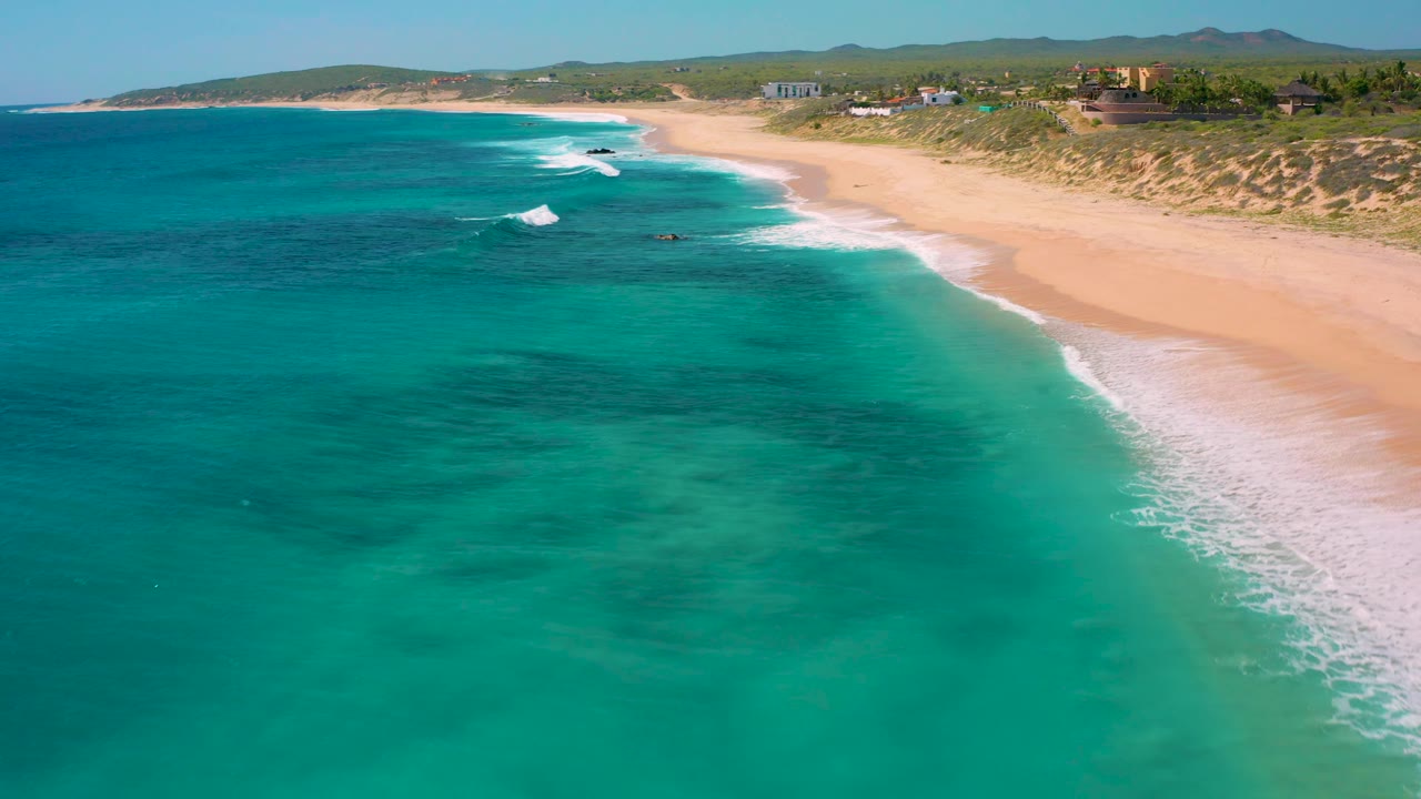 Aerial view of El Cardoncito with blue-green shallows filling most of the frame, white surf touching the beach at right, scattered houses along the dunes, and no visible people.