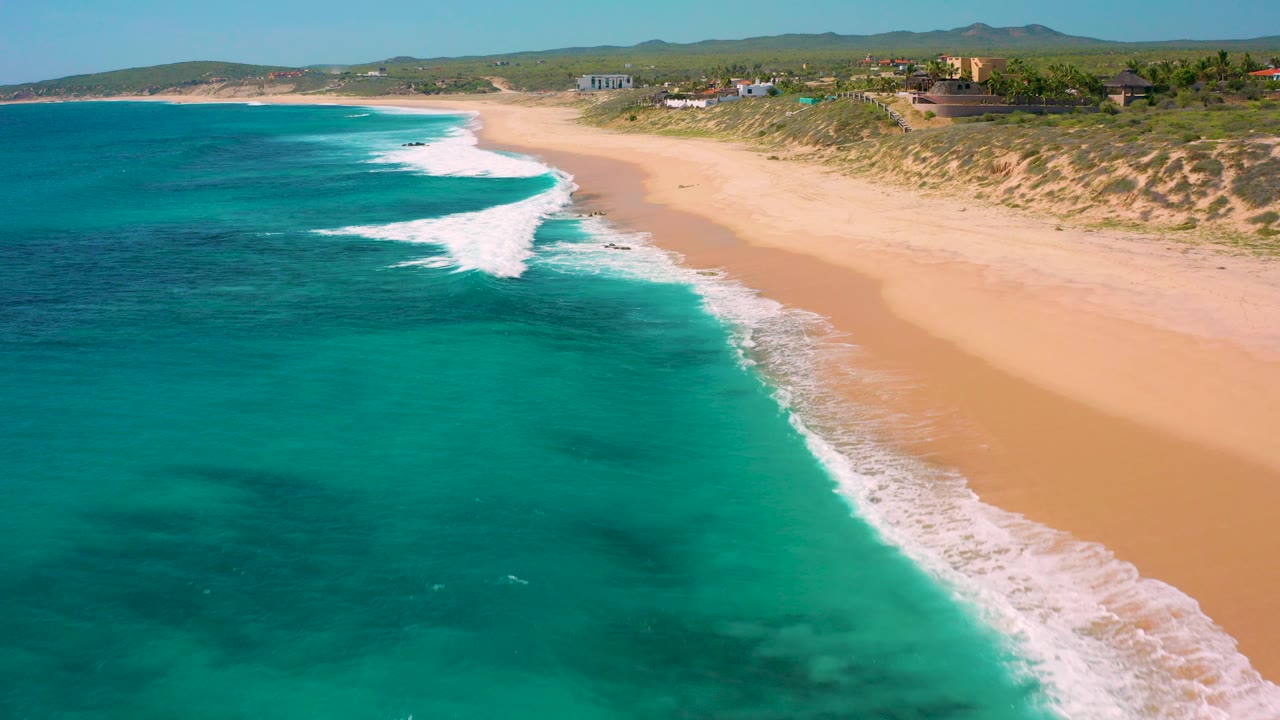 Aerial frame over El Cardoncito with turquoise water in the foreground, a diagonal white wave line reaching sand, dunes and distant buildings beyond the beach, and no visible people.