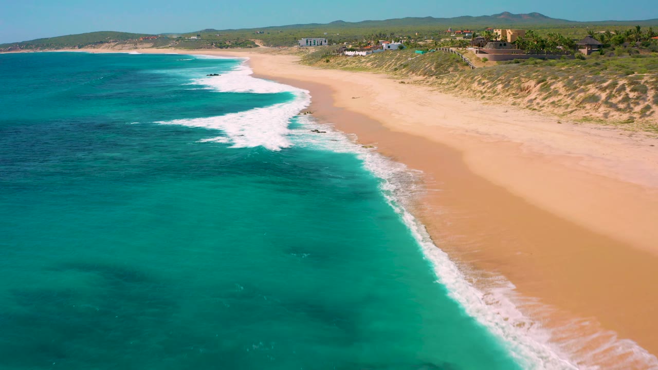 Aerial view in El Cardoncito where a curved white breaker spreads toward the sand, darker patches show through the water, dunes and small shoreline structures sit in the background, and no visible people appear.