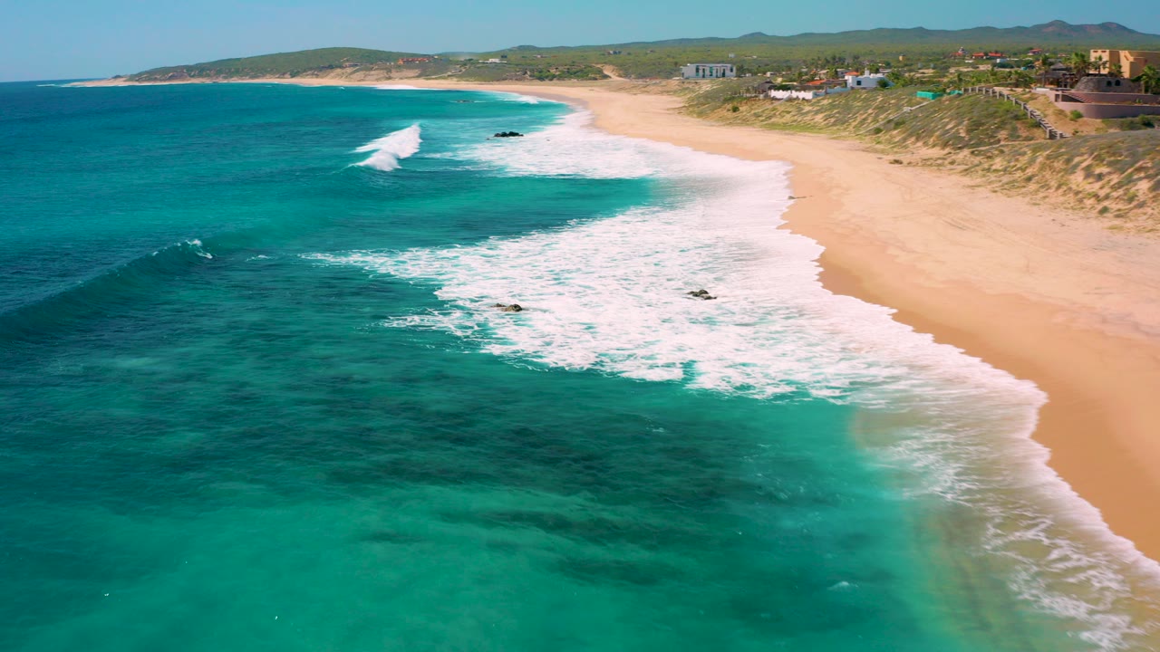 Aerial view over El Cardoncito coastline in Baja California Sur with turquoise water in the foreground, white surf along a sandy beach, low inland hills, and no visible people.