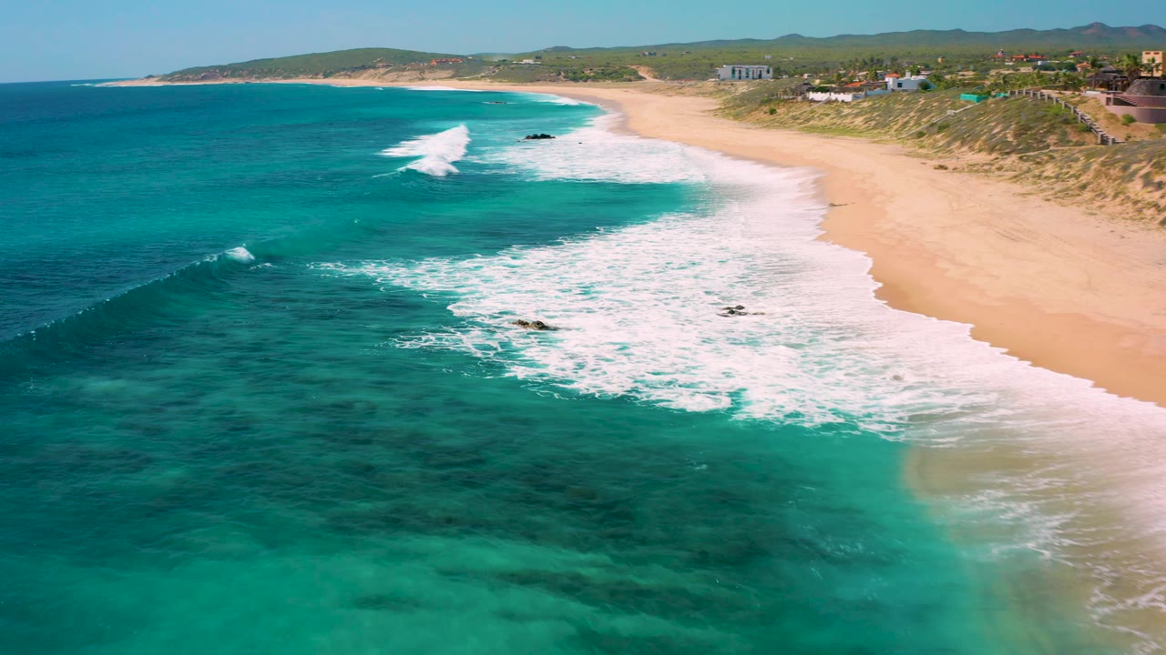 Aerial frame from El Cardoncito showing turquoise sea on the left, a curved band of white surf, empty sandy beach on the right, low vegetation behind the dunes, and no visible people.