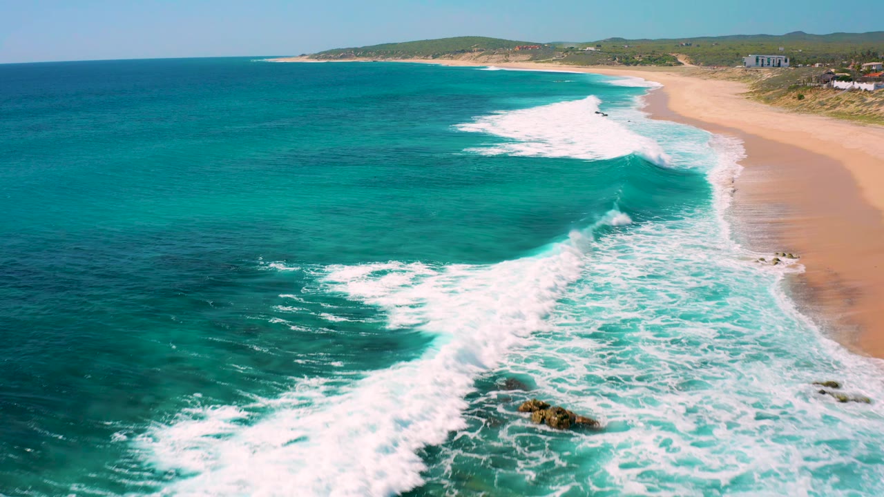 Aerial view at El Cardoncito, Baja California Sur with a broad white wave band crossing turquoise water toward the empty sand, with no visible people.