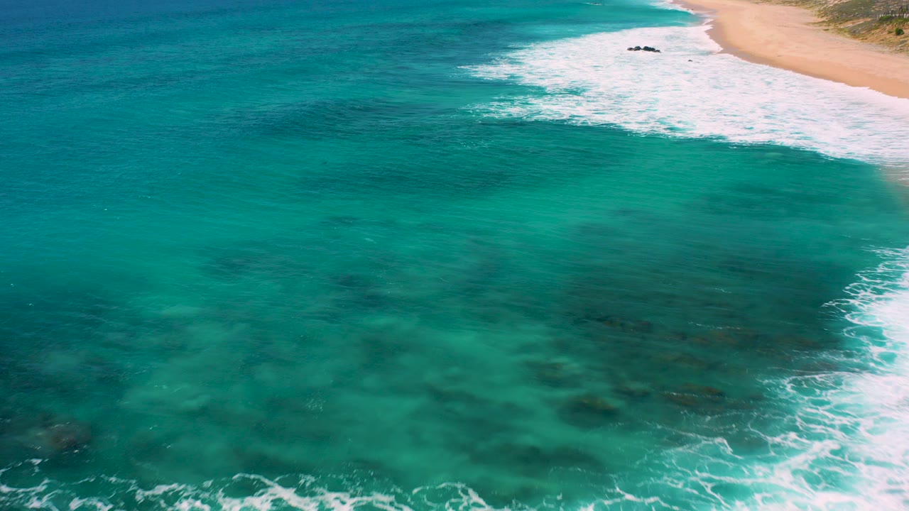 Aerial view over clear green water at El Cardoncito, Baja California Sur, with white surf curling beside a sandy beach and no visible people.