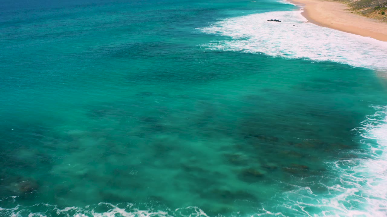Clear green ocean fills most of the frame at El Cardoncito, with white foam pushing diagonally toward the sandy shoreline and no visible people.
