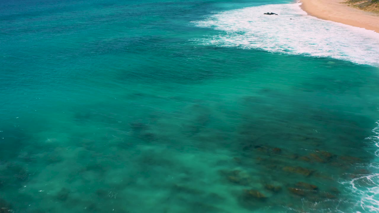 Aerial frame from El Cardoncito showing turquoise-green water, darker submerged areas, white surf, and a sandy beach strip with no visible people.