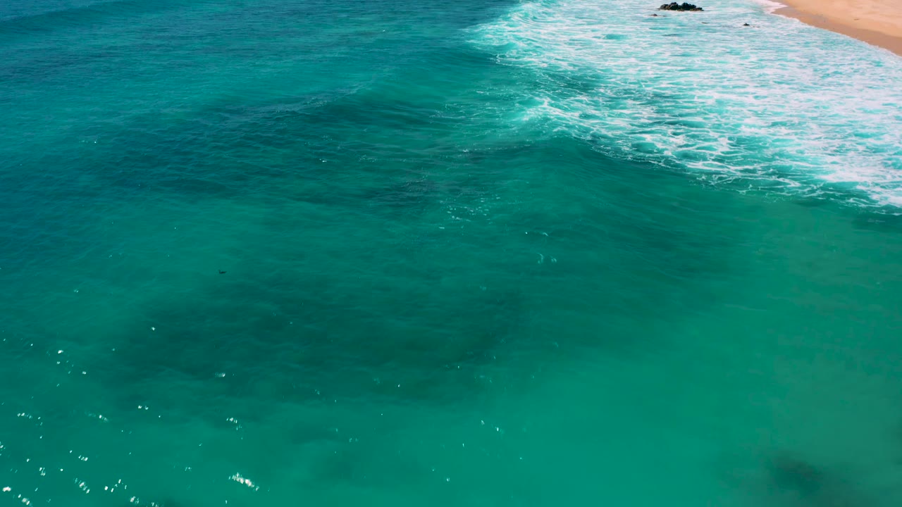 Aerial frame over clear green-blue water at El Cardoncito, with foamy surf crossing the upper frame and no visible people.