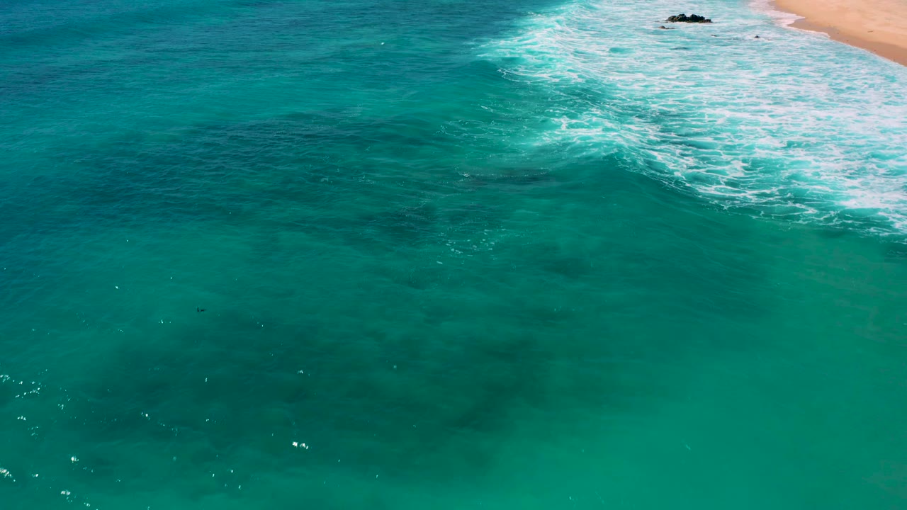 High angle view of turquoise ocean off El Cardoncito, with white breakers near the sandy shore and darker areas under the water, no visible people.