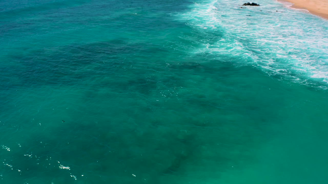 Aerial coastal view at El Cardoncito with surf foam spreading diagonally across clear water and a narrow sandy edge visible, no visible people.