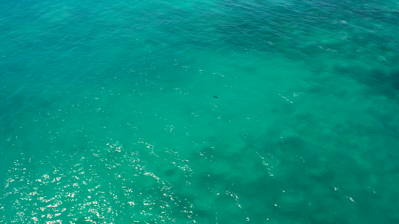 Aerial view over turquoise water in El Cardoncito, Baja California Sur, with darker underwater shapes, bright surface glints, and no visible people.