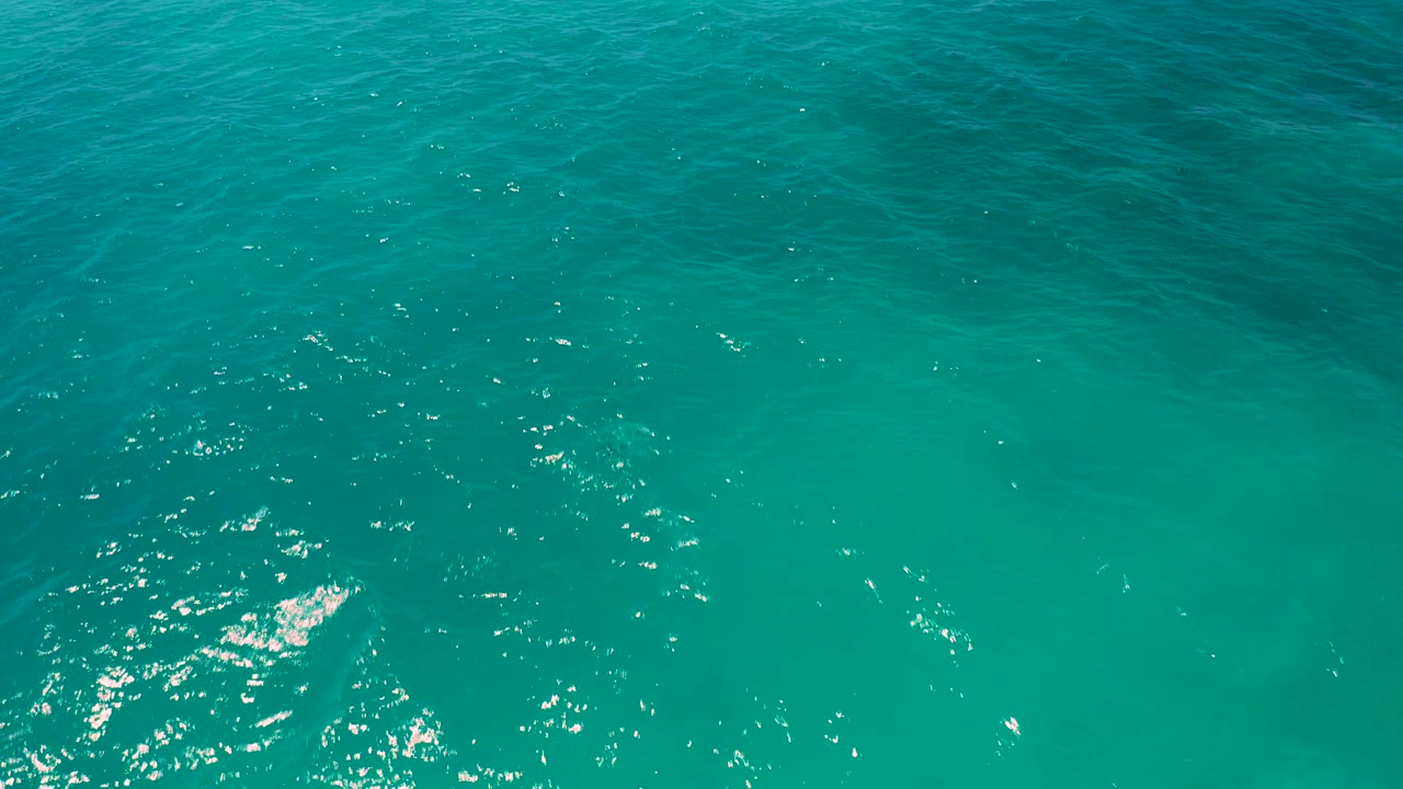 Aerial frame of El Cardoncito water with bright sparkles, gentle surface chop, blue-green color shifts, a tiny dark fish-like object near the top, and no visible people.