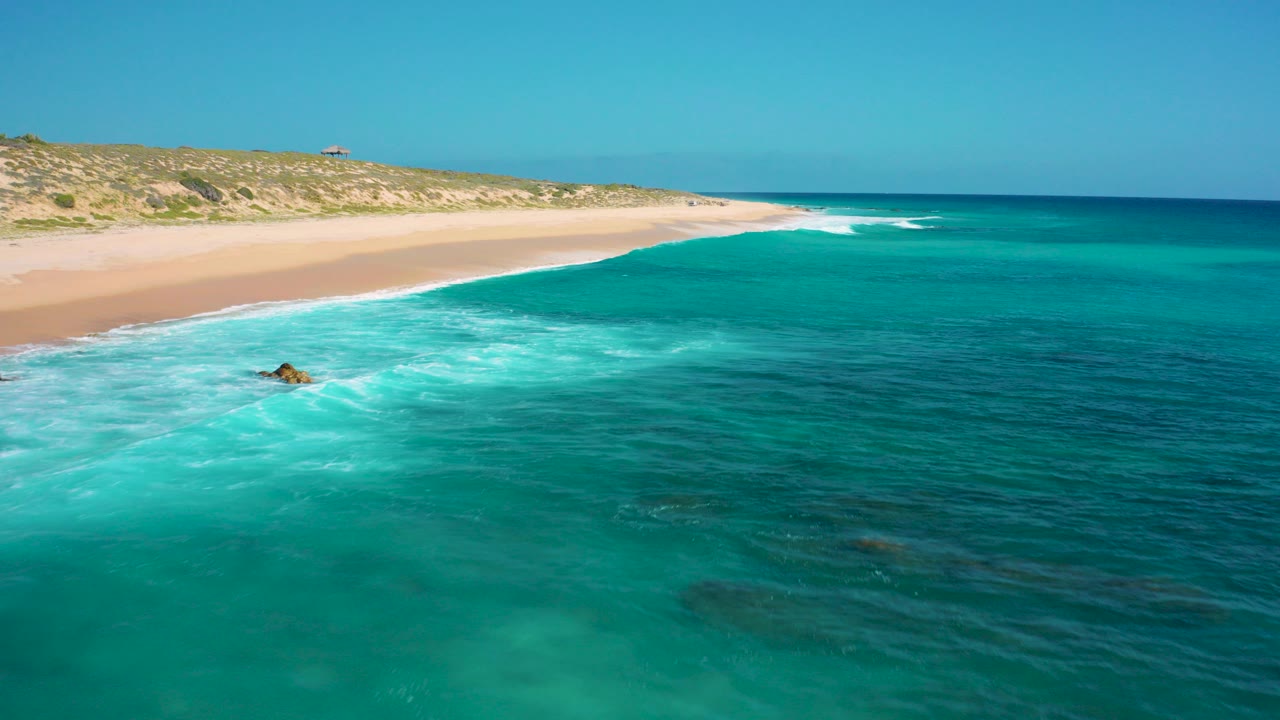Aerial frame over El Cardoncito, Baja California Sur, showing turquoise water, a pale sandy beach, low dunes, a small offshore rock, and no visible people.