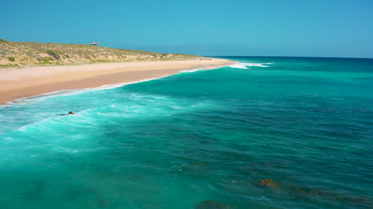 Drone frame from El Cardoncito showing a longer stretch of beach at left, turquoise shallows across the center, darker water offshore, and no visible people.