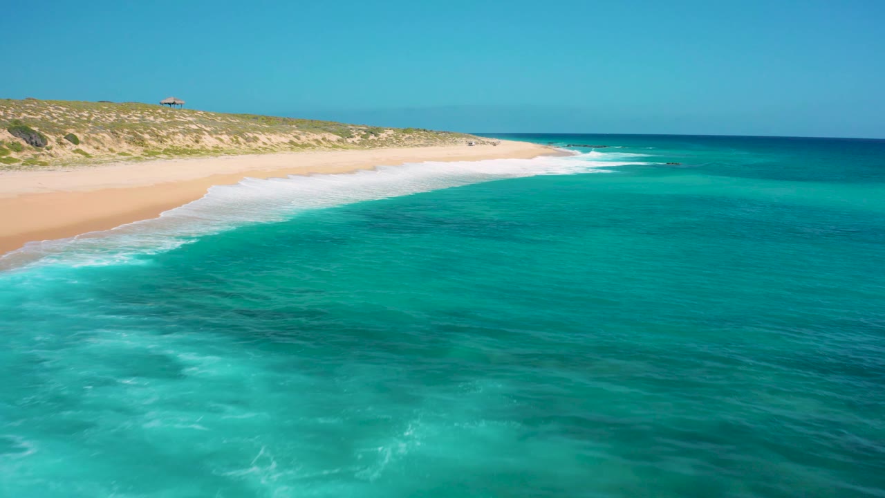 A wide aerial view of El Cardoncito shows shallow turquoise water in front, a curved sandy shoreline, scrub-covered dunes, and no visible people.