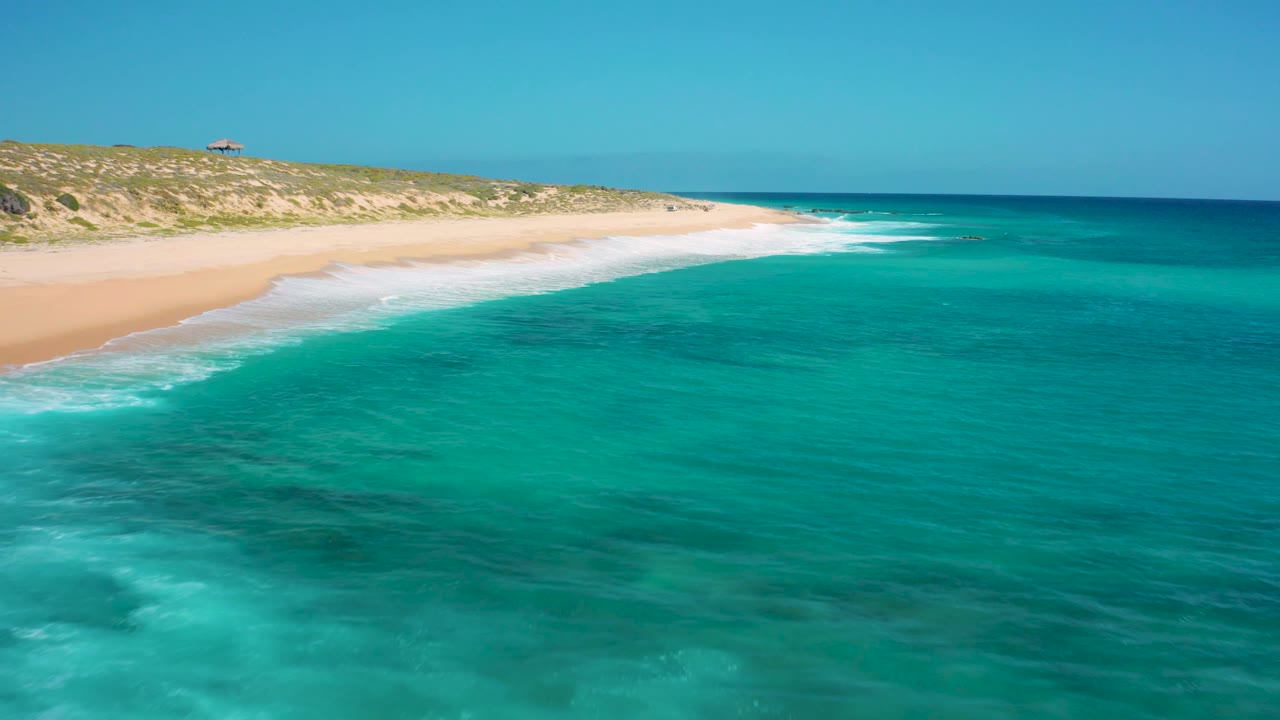At 0:03 the aerial composition holds the long El Cardoncito beach along the left, calm blue-green sea across the right, and no visible people.
