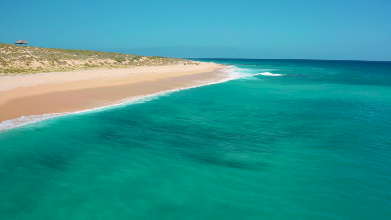 Wide aerial frame from El Cardoncito with turquoise sea in front, a pale sand beach sweeping toward the center, low green dunes behind the beach, and no visible people.