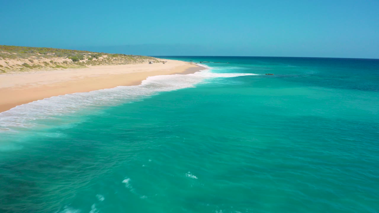 Aerial frame over El Cardoncito with turquoise water in the foreground, pale sand curving left, low dune vegetation behind the beach, and no visible people.