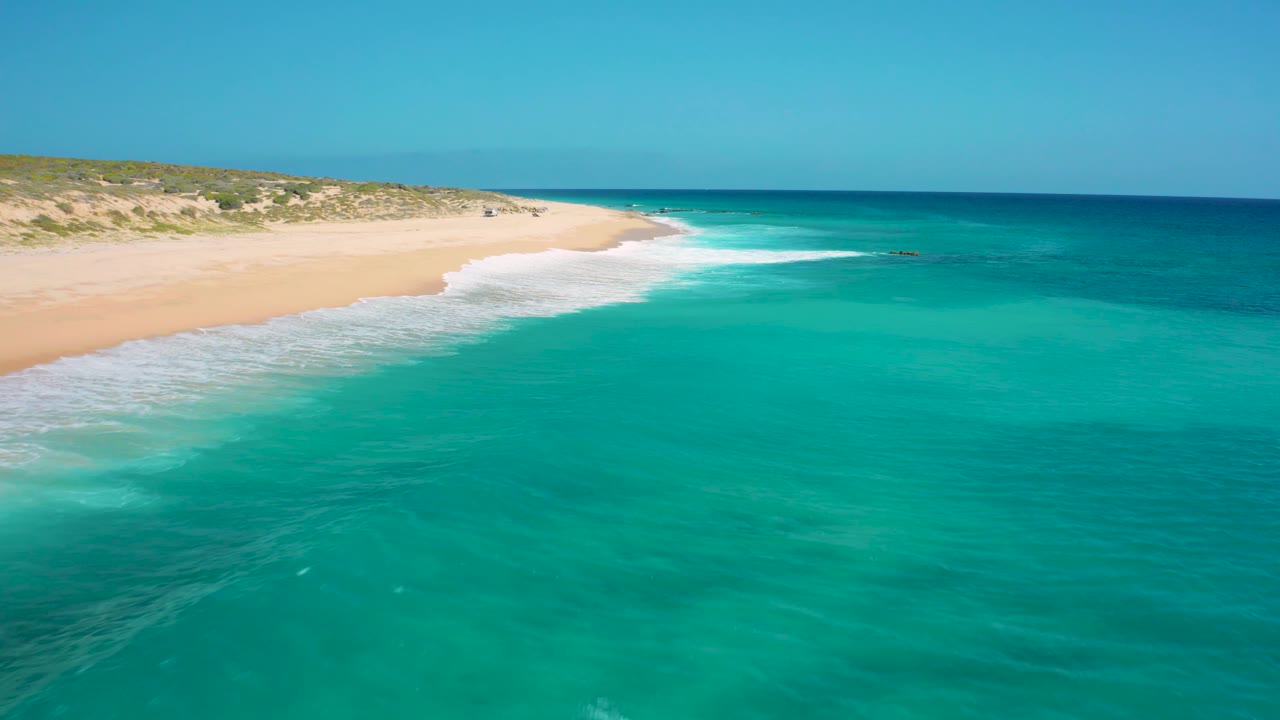 Aerial view in El Cardoncito with more open turquoise water in the foreground, a curving sandy beach at left, darker blue sea near the horizon, and no visible people.