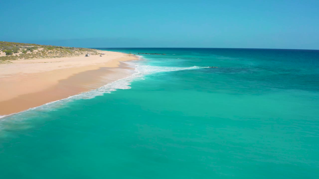 Aerial frame at El Cardoncito showing blue-green water curving into a sandy shoreline, scattered rocks near the surf, and no visible people.