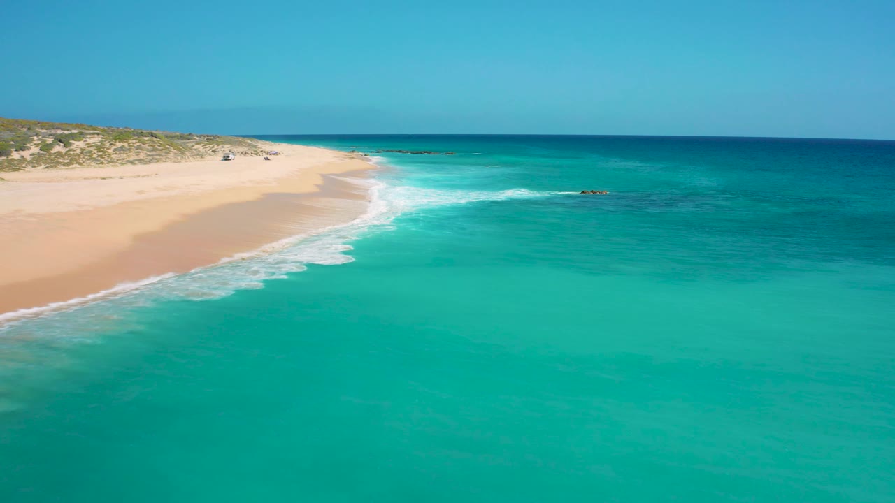 Wide aerial view at El Cardoncito with clear turquoise water in front, sandy beach along the left, low dunes behind it, and no visible people.