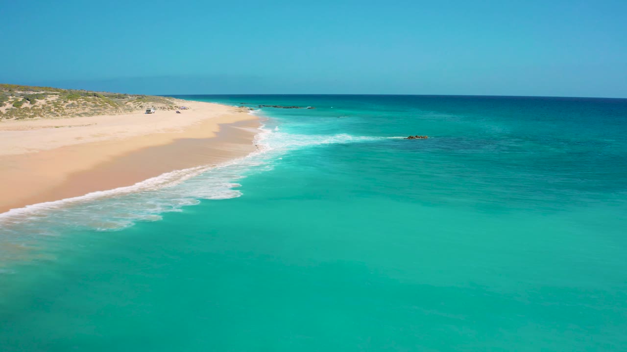 Aerial frame at El Cardoncito with a foamy wave breaking onto pale sand, shallow turquoise water in the foreground, and no visible people.