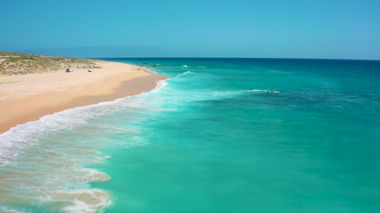 Aerial view at El Cardoncito, Baja California Sur, with green-blue water filling the foreground, a sandy beach on the left, low dunes in back, and no visible people.