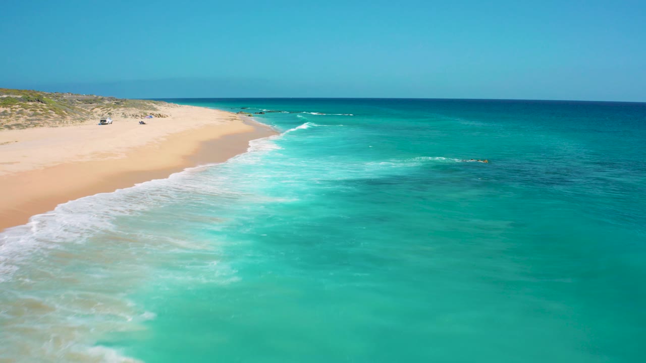 The El Cardoncito shoreline runs diagonally through the frame, with aquamarine water to the right, white wash on the sand, low dunes at upper left, and no visible people.