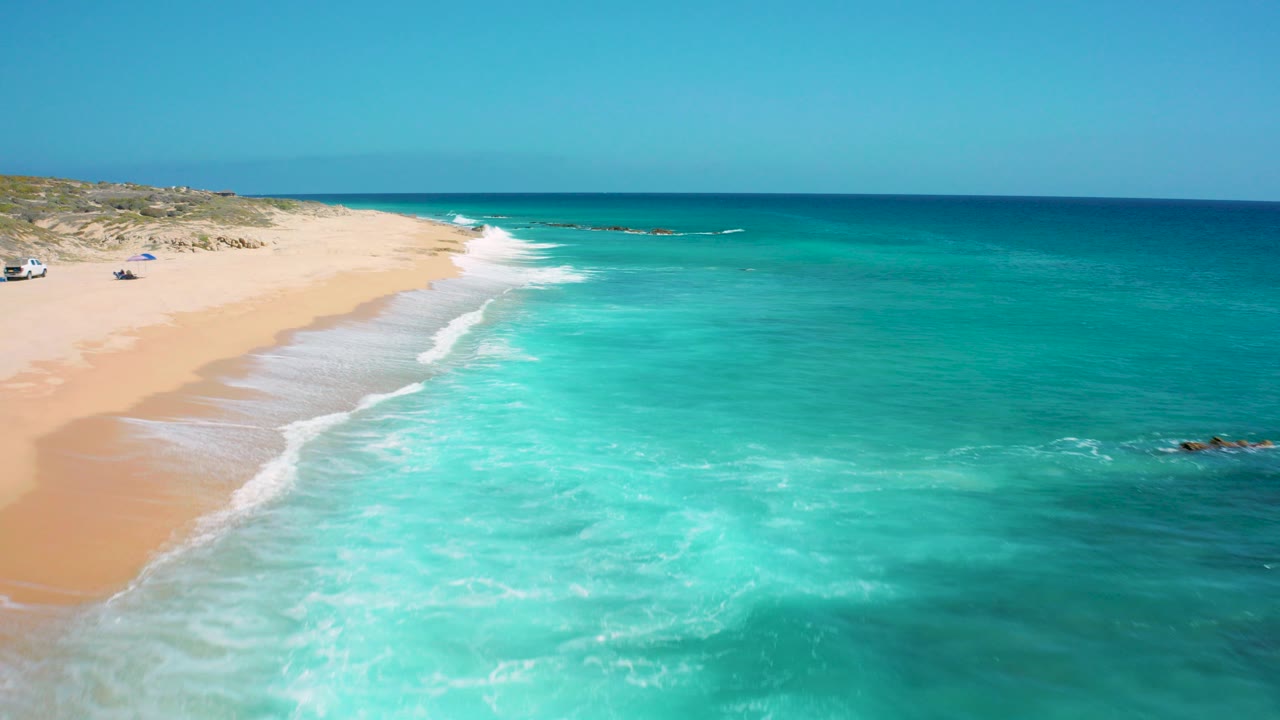 Aerial view over El Cardoncito, Baja California Sur, with turquoise surf spreading toward a sandy beach, low dunes at left, deep blue water beyond, and no visible people.