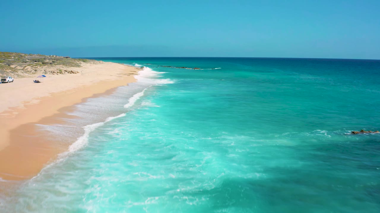 Aerial frame of El Cardoncito shoreline with white foam bands crossing shallow green water, open sand on the left, darker ocean on the right, and no visible people.
