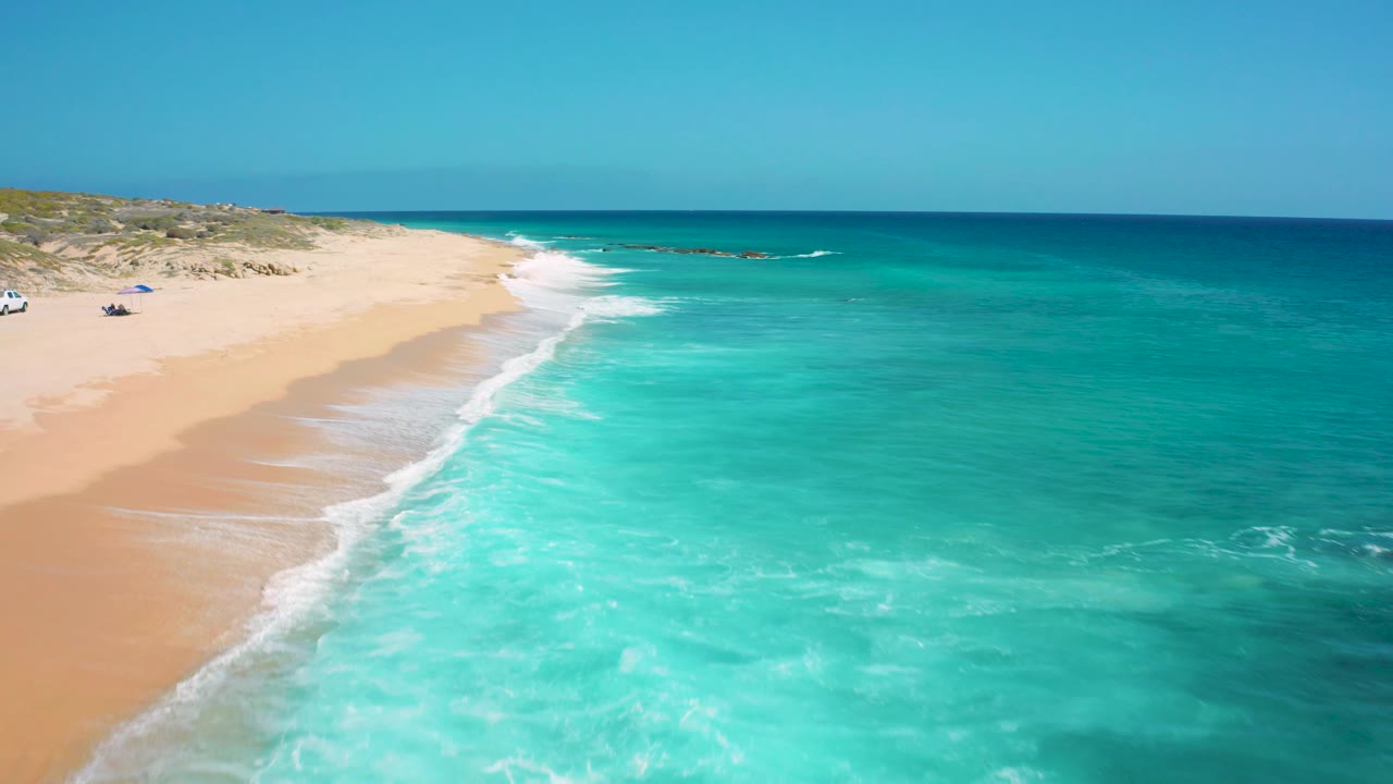 Aerial view of El Cardoncito with the sandy shoreline curving away, bright turquoise surf in the foreground, darker blue sea near the horizon, and no visible people.